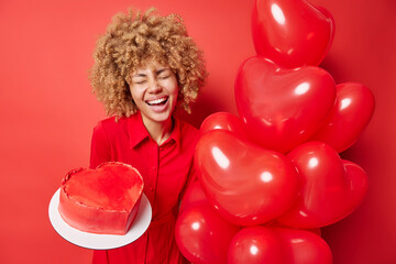 Happy Valentines Day. Cheerful woman with curly hair wears dress holds heart inflated balloons and cake celebrates special occasion isolated over red background. Overjoyed female model being on party