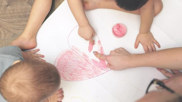 baby children drawing with finger paints pink heart white paper. mother teaching kids toddler painting using hand paint. early education home. unrecognizable child sitting on floor indoors coloring