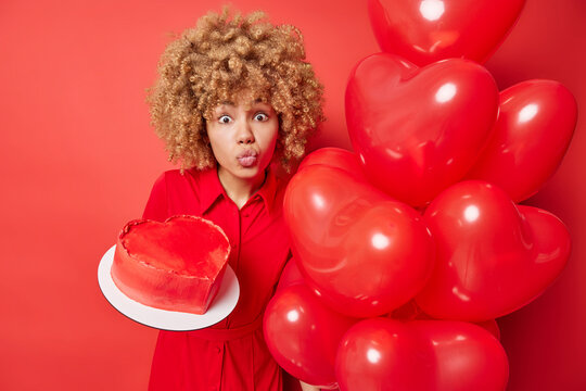 Romantic curly haired woman keeps lipd folded holds delicious cake in form of heart and inflated balloons wears dress isolated over vivid red background. People holidays celebration concept.