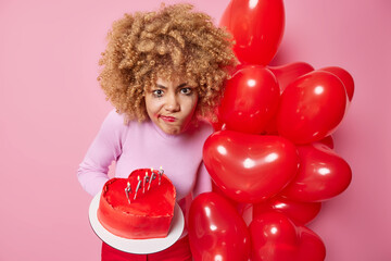 Indoor shot of frustrated curly haired woman purses lips feels displeased celebrates Valentines Day alone holds heart cake and inflated red balloons isolated over pink background feels very angry