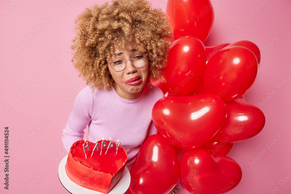 Wall mural upset young woman with curly hair has spoiled makeup after crying holds heart sweet cake and bunch o