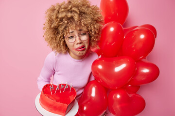 Upset young woman with curly hair has spoiled makeup after crying holds heart sweet cake and bunch of inflated balloons wears spectacles and casual jumper celebrates festive event poses indoor