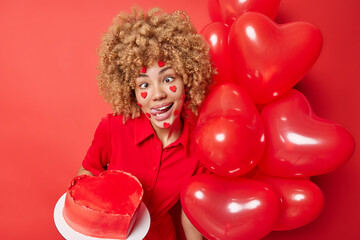 Funny curly haired young woman foolishes around makes grimace holds heart balloons and cake poses against vivid red background entertains someone celebrates birthday Lovers Day. Holidays concept