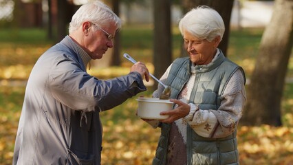 Kind caucasian retired grey-haired man helps people in need by sharing his homemade soup with elderly homeless woman. High quality photo