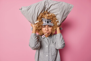 Dissatisfied curly haired woman keeps pillow over head wears sleepmask and pajama prepares for sleep isolated over pink background keeps wooden toothbrush in mouth. Sleeping rest and bedtime