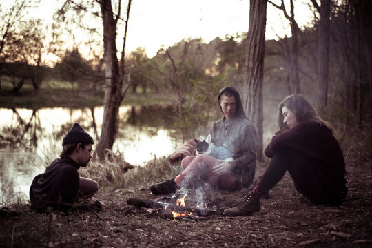 friends sit contemplating by river and campfire with dogs on lap