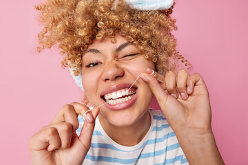 Close up shot of young cheerful woman uses tooth floss for cleaning teeth winks eyes has curly fair...