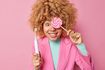 Positive curly haired woman covers eye with delicious candy holds electric toothbrush has sweet tooth dressed in elegant clothes isolated over pink background. Teeth problem and harmful food