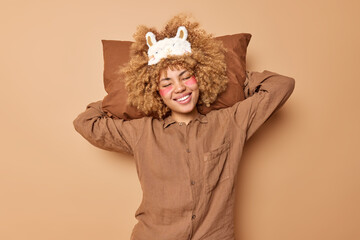 Pleased young curly woman keeps soft pillow behind head applies hydrogel patches under eyes dressed...
