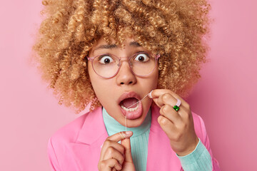 Close up portrait of shocked young curly haired woman flosses her teeth stares bugged eyes has...