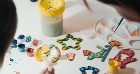 Child with grandmother paints homemade Christmas tree toys made of wood for Christmas, close-up shot, rear view. Concept of development, joint family pastime and upbringing of child in playful way