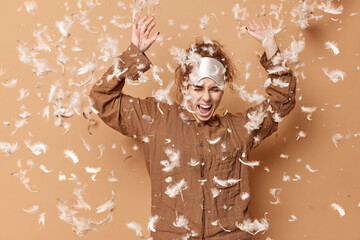 Overjoyed carefree young woman has fun wears sleepmask on forehead and pajama exclaims happily poses against beige background with feathers flying in air after pillow fight. Awakening concept