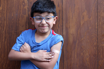 Little 6-year-old Latino boy with glasses and a blue shirt shows his arm with a bandage because he...