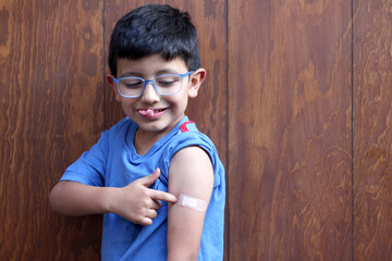 Little 6-year-old Latino boy with glasses and a blue shirt shows his arm with a bandage because he...