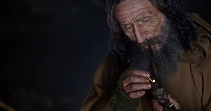Fantastic monk sorcerer with a gray beard chids in a cave and lights a pipe with tobacco. Close up.