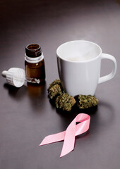 cancer pink ribbon with marijuana buds, white cup with infusion steam, oil drops dispenser bottle, on brown table, wooden background