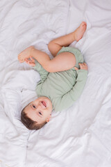 Fototapeta na wymiar cute smiling baby in a green cotton bodysuit is lying on a white bed on his back. top view