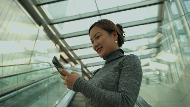 Charming Adult Chinese Woman Playing Cellphone Standing on Escalator Casually, Checking Messages on Smart Phone with Smile in Chengdu