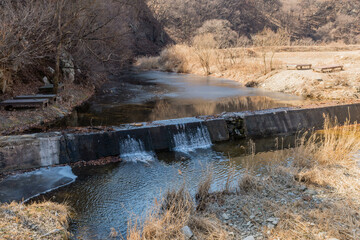 Obraz premium River water flowing over concrete spillway on sunny winter day.