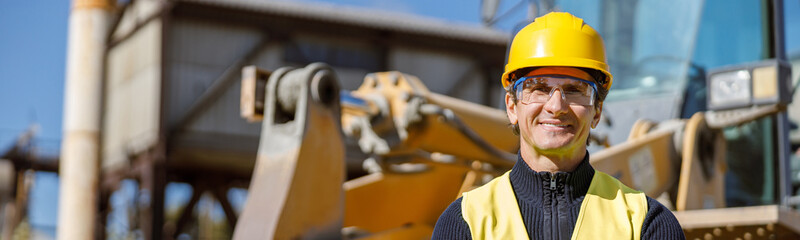 Joyful matured man engineer wearing safety helmet, glasses and vest while keeping arms crossed and...
