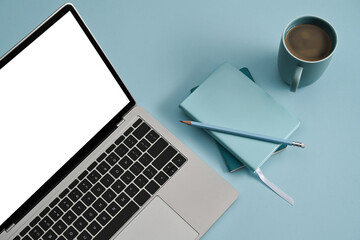 Mockup computer laptop with white screen, notebooks and coffee cup on blue background.