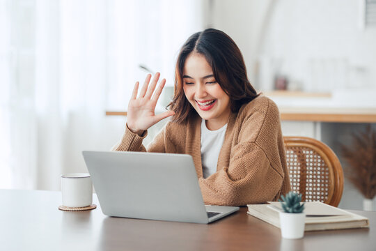 Happy positive young asian woman enjoying online communication at home, Female using wifi while video conferencing with friend, sitting in front of open laptop, smiling and waving hand, saying hi