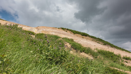 A hillside against a cloudy sky. Sulfurous deposits from fumaroles are visible on the soil. In the...