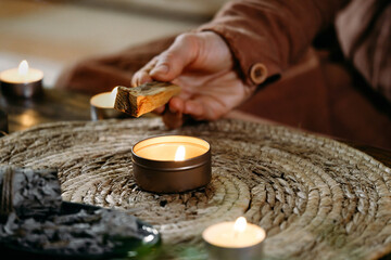 Woman hands burning Palo Santo, before ritual on the table with candles and green plants. Smoke of smudging treats pain and stress, clears negative energy and meditation wooden stick