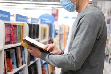 A man in a medical mask looks at a book in the library. Distance learning. Buying a book in a bookstore.