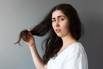 Portrait of young upset woman show dry tips of her long dark hair. Gray background. The concept of trichotillomania