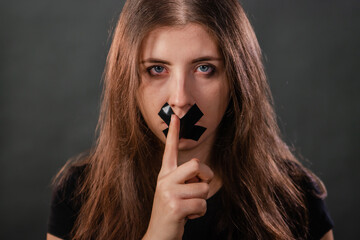 Obraz premium Portrait of a young woman with her mouth sealed with black tape, restriction of freedom of speech, censorship and a ban on speaking and expressing her opinion, isolated on a dark background.