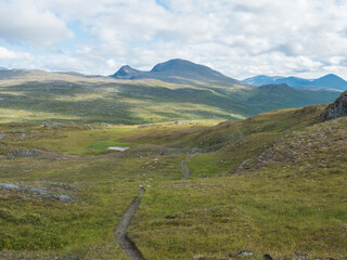 Fototapeta na wymiar Footpath in northern artic landscape, tundra in Swedish Lapland with green hills and mountains at Padjelantaleden hiking trail. Summer day, blue sky, white clouds