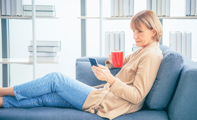 Obraz premium Happy beautiful relaxed mature older woman drinking coffee relaxing on sofa at living room. Elderly woman lying on sofa using smartphone and drinking coffee at home. 
