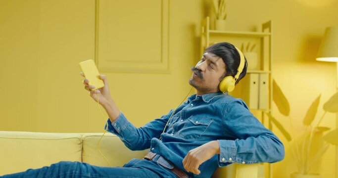 Happy hispanic man with mustache listens to favorite music using a mobile phone and headphones while imagining playing a musical instrument on the sofa in living room with monochrome yellow color room