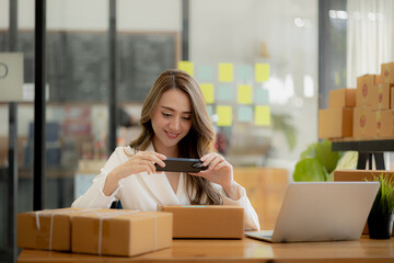 A woman using a smartphone to take pictures in front of parcel boxes, parcel boxes for packing goods, delivering goods through private courier companies. Online selling and online shopping concepts.