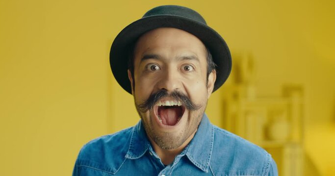 Headshot of amazed hispanic young man with a mustache and wear a head looking at camera with open mouth while standing against yellow background in the studio. Man looking with amazed expression