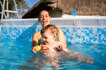 Fototapeta premium Mother laughing while playing with her baby inside a pool in summer