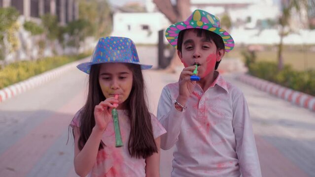 Indian kids wearing colorful hats having fun blowing blow-out whistles together - Holi celebrations  childhood game  leisure time. Cheerful siblings celebrating Holi - family bonding  young brother...