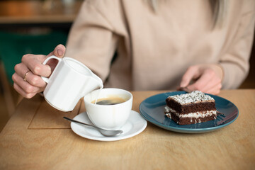 Cropped photo of blurred woman pouring milk cream in coffee, eating brownie cake. Cafe holiday pleasure, close up