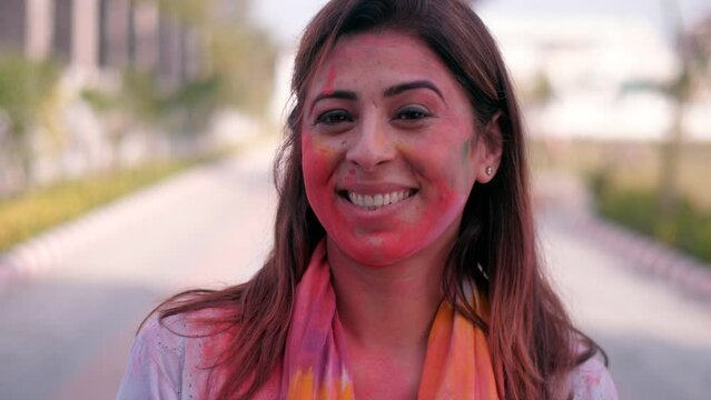 A beautiful Indian lady with her face covered in Holi colors smiling happily for the camera - a toothy smile  a Hindu festival. A pretty female enjoying the festival of colors in a public park - a ...
