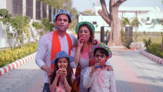 A happy Indian family with their faces smeared in Holi colors blowing blow-out whistles - Holi celebrations  childhood games  leisure time. A young couple and little kids celebrating Holi in a publ...