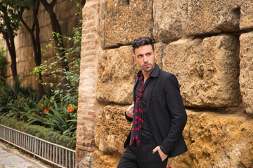 Fototapeta na wymiar Portrait of young Spanish man, wearing black shirt, jacket and pants, with black handkerchief with red polka dots, posing next to a stone wall. Flamenco concept, art, dance, culture, tradition.
