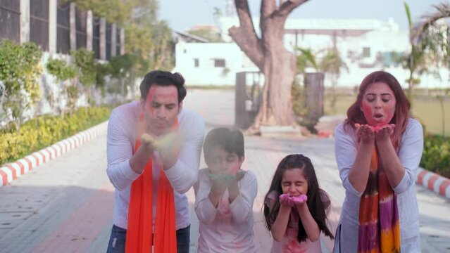 A modern nuclear family celebrating Holi on a farm - Holi party. An urban Indian family with their faces smeared with Holi colors blowing off Abeer from their hands - a safe Holi  vibrant colors  f...