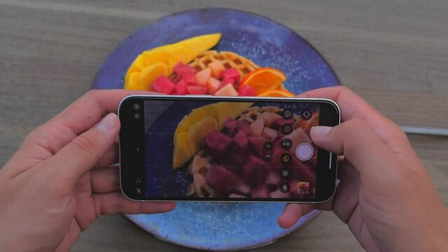 Female hands using smartphone to take photos of tasty waffle with red fruits and mango