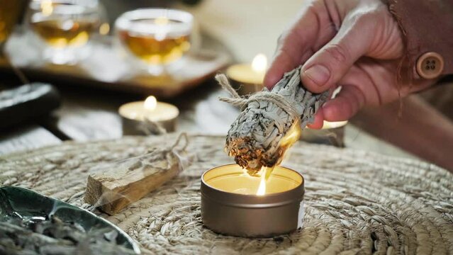 Woman hands burning white sage, before ritual on the table with candles and green plants. Smoke of smudging treats pain and stress, clear negative energy and meditation