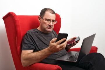 Middle-aged man in glasses with a laptop on his lap and telephones sitting in a red armchair. mockup