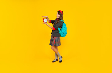 happy kid in school uniform and backpack hold alarm clock on yellow background, student