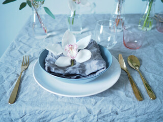 Colorful table setting. Grey linen tablecloth and napkin, golden cutlery and colorful tableware. Orchid flower on top of a napkin. Table setting with flowers.