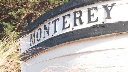 Obraz premium Monterey sign on white wooden boat stern, fisherman nautical vessel stem, bow or rostrum near Cannery Row and Bay Aquarium. Coniferous pine cypress tree. Historic capital, tourist city, California USA