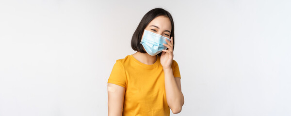 Covid-19, vaccination and healthcare concept. Cute asian girl in medical face mask, showing band...